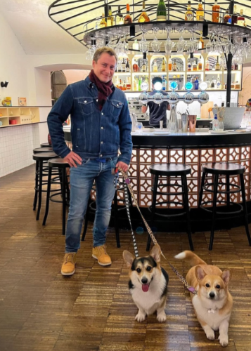 Egy mosolygó férfi farmer ruhában, sálban áll egy kávézóban vagy bárban, pórázon tartva két aranyos corgi kutyát, akik előtte ülnek a földön. Férfi, corgi, kutya, kávézó, állatbarát hely, szabadidő, kutyasétáltatás, barátok, állatokkal az élet.