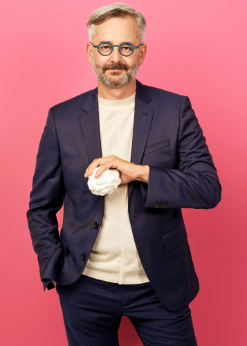 Szemüveges, őszes hajú férfi sötétkék zakóban és fehér pulóverben, élénk rózsaszín háttér előtt egy fehér, rücskös gömböt tart a kezében. Üzleti portré, kreatív szakember, stúdiófotó, modern elegancia, kék zakó, rózsaszín háttér.